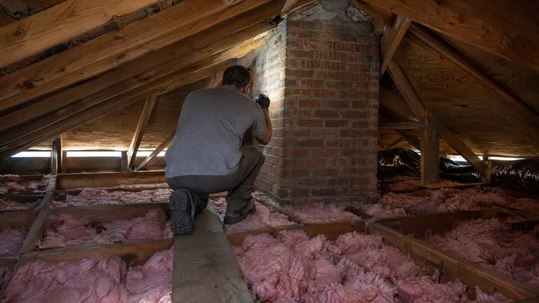 A technician in an attic shines a flashlight along a brick chimney where it passes the roof framing.