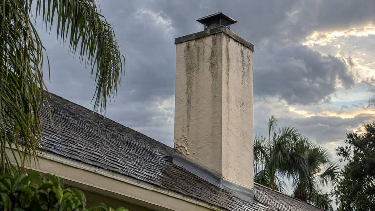 A cream stucco chimney with dark rain streaks and peeling paint near its base on a wet shingle roof.